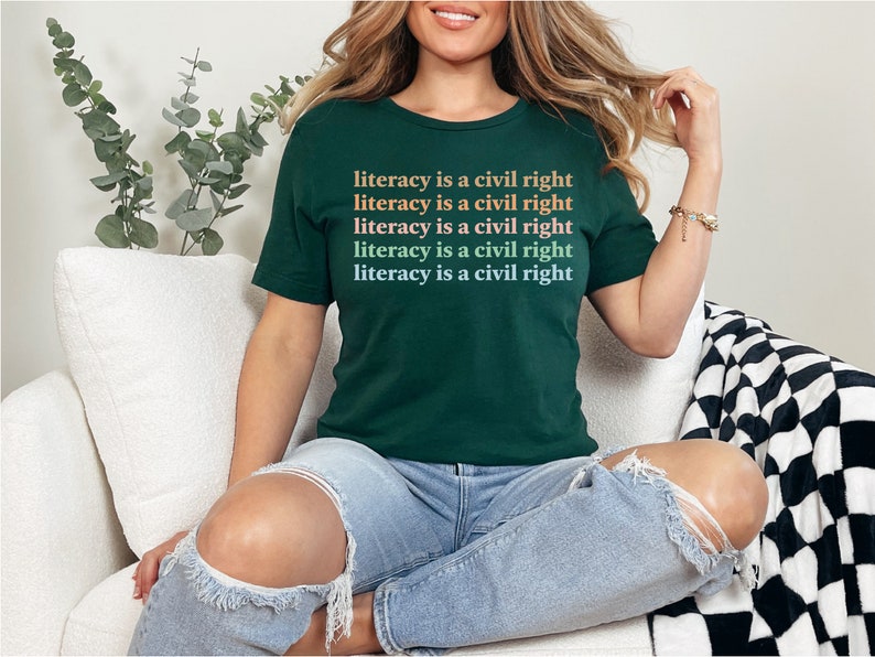 May include: A woman wearing a dark green t-shirt with the text "literacy is a civil right" repeated five times in a gradient of orange, pink, and blue. She is sitting on a white sofa with a black and white checked throw blanket.
