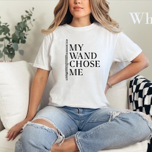 May include: A white short-sleeved t-shirt featuring a black graphic of a flute and the text "MY WAND CHOSE ME" printed on the front. The shirt has a crew neckline and a standard fit, showcasing a musical instrument design.