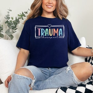 May include: A navy blue t-shirt with a white frame and the text "Trauma therapist" in rainbow colors. A small heart is above the text.