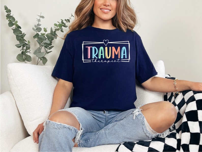 May include: A navy blue t-shirt with a white frame and the text "Trauma therapist" in rainbow colors. A small heart is above the text.