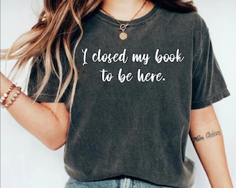 May include: Dark gray t-shirt with the white text "I closed my book to be here." The shirt is a crew neck style and appears to be made of a soft, comfortable material. The shirt is worn with blue jeans.