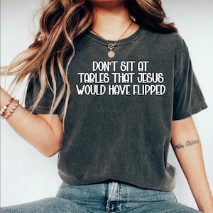 Peut inclure: T-shirt gris foncé avec le texte blanc "DON'T SIT AT TABLES THAT JESUS WOULD HAVE FLIPPED." Le t-shirt a des manches courtes et un col rond. Le modèle porte un jean bleu.