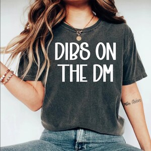 May include: Dark gray t-shirt with the white text "DIBS ON THE DM". The shirt is a casual style, suitable for everyday wear. The model is wearing blue jeans.