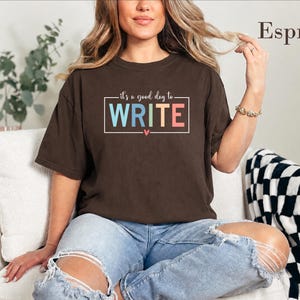 May include: A dark brown short-sleeve t-shirt featuring the text "it's a good day to WRITE" in a rectangular frame with a small heart icon. The word WRITE is printed in large, multi-colored block letters, and the word Espresso is visible.