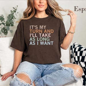 May include: A dark brown short-sleeve t-shirt featuring the text "IT'S MY TURN AND I'LL TAKE AS LONG AS I WANT" in stacked lines of orange, pink, green, and blue lettering. The word "Espresso" is printed in the upper right corner.
