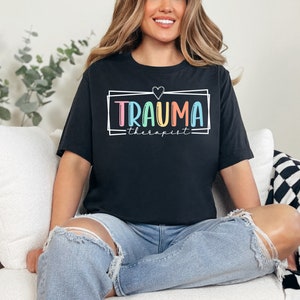 May include: A black t-shirt with a white frame around the text "Trauma Therapist" in rainbow colors. A small heart is above the text.