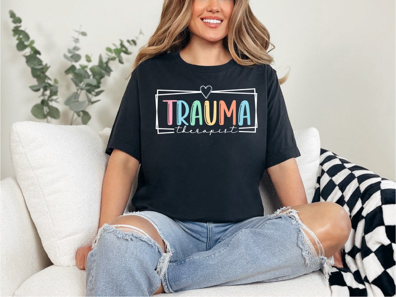 May include: A black t-shirt with a white frame around the text "Trauma Therapist" in rainbow colors. A small heart is above the text.