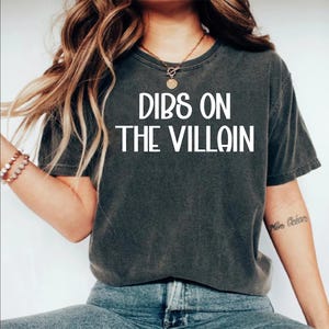 May include: Dark gray t-shirt with the text "DIBS ON THE VILLAIN" in white block letters. The shirt is a casual style, likely made of cotton or a similar fabric. The model is wearing blue jeans.