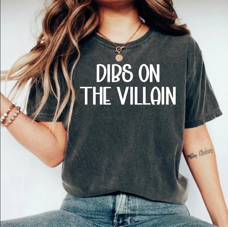 May include: Dark gray t-shirt with the text "DIBS ON THE VILLAIN" in white block letters. The shirt is a casual style, likely made of cotton or a similar fabric. The model is wearing blue jeans.