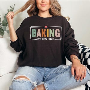 May include: A black sweatshirt with the text "Baking - It's how I hug" in a colorful box with a small red heart above the text.
