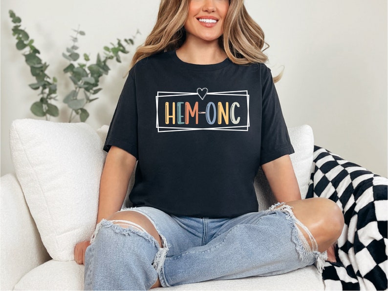 Puede incluir: Una camiseta negra con un contorno blanco de un coraz&oacute;n y un cuadro alrededor del texto "HEM-ONC" en colores arco&iacute;ris.