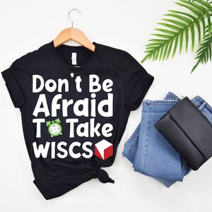May include: Black t-shirt with white text that reads "Don't Be Afraid To Take WISC-S". A green alarm clock and a red cube are next to the text.
