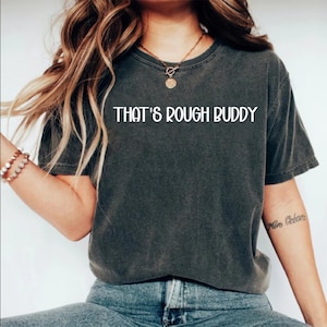 May include: Dark gray t-shirt with the text "THAT'S ROUGH BUDDY" in white. The shirt has short sleeves and a crew neck. The shirt is made of a soft, comfortable material.