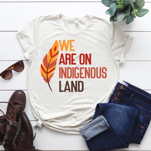 May include: A beige t-shirt with the text "WE ARE ON INDIGENOUS LAND" in orange, red, and yellow lettering. A stylized orange and red feather is to the left of the text.