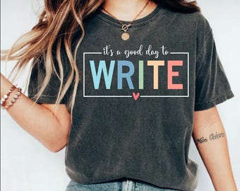 Może przedstawiać: Szary t-shirt z napisem "it's a good day to WRITE" w kolorowej czcionce tęczowej. Tekst jest zamknięty w białym prostokącie z małym różowym sercem poniżej.