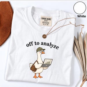 May include: A white short-sleeve t-shirt featuring a graphic of a goose wearing a green cap and glasses, carrying a brown satchel, and holding a laptop displaying a chart. The text "off to analyze" is printed above the goose in black.