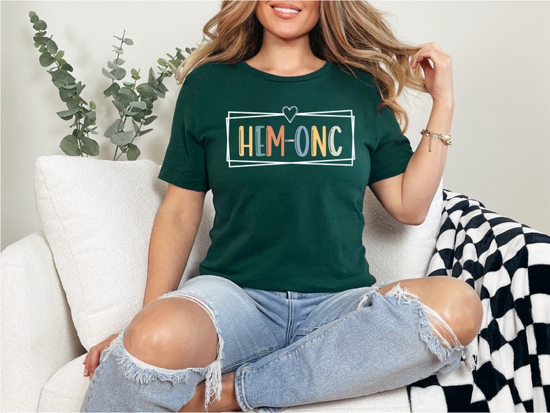 Puede incluir: Una mujer lleva una camiseta verde oscuro con el texto "HEM-ONC" en letras de colores dentro de un marco blanco. La camiseta tiene un peque&ntilde;o coraz&oacute;n sobre el texto. Est&aacute; sentada en un sof&aacute; blanco con una manta de cuadros blancos y negros.