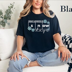 May include: A black short-sleeve t-shirt featuring the white text "PROUD SUPPORTER OF" above a grid containing the letters "s n ow" and the word "days" in script, accented with light blue snowflake graphics and the word "Black" in the corner.