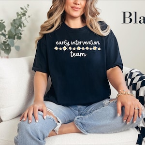 May include: A black short-sleeved t-shirt featuring the white text "early intervention team" centered on the chest, accented by a horizontal row of small white and yellow daisy flowers positioned between the two lines of text.