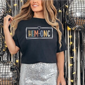 Puede incluir: Una camiseta negra con un gr&aacute;fico de colores arco&iacute;ris que dice "HEM-ONC" con un coraz&oacute;n blanco sobre el texto.