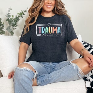 May include: A dark grey t-shirt with the text "TRAUMA therapist" in a colorful rainbow font inside a white outlined rectangle with a small white heart above the text.
