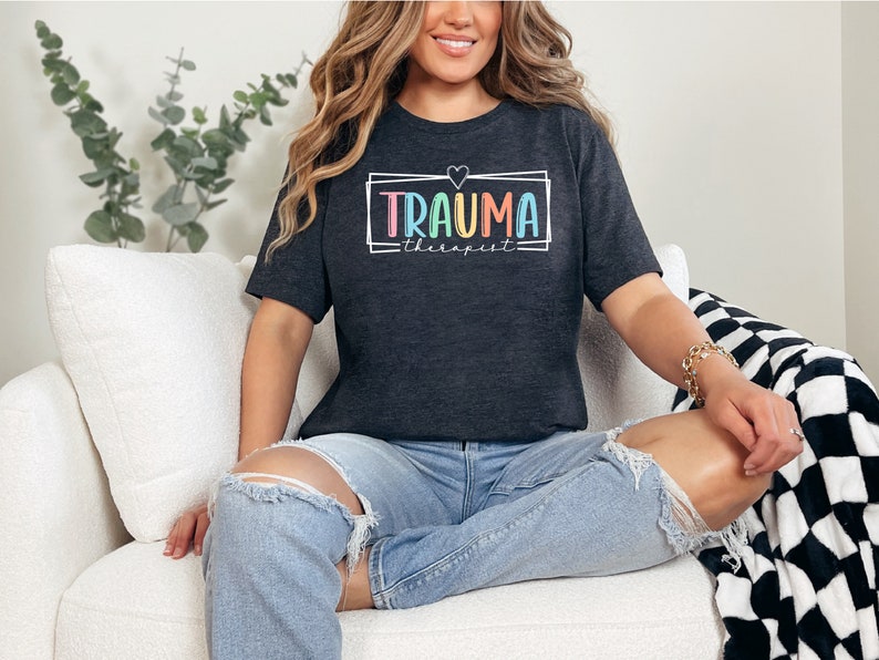 May include: A dark grey t-shirt with the text "TRAUMA therapist" in a colorful rainbow font inside a white outlined rectangle with a small white heart above the text.