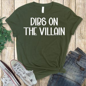 May include: An olive green t-shirt with the text "DIBS ON THE VILLAIN" in white block letters. The shirt is paired with blue jeans and white sneakers with red accents.