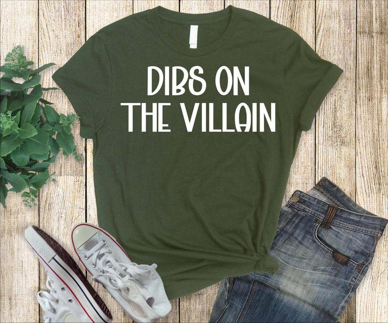 May include: An olive green t-shirt with the text "DIBS ON THE VILLAIN" in white block letters. The shirt is paired with blue jeans and white sneakers with red accents.