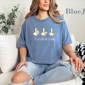 May include: A blue jean colored t-shirt featuring a graphic of three white geese holding baking items with the text "IT'S A GOOD DAY TO BAKE" printed below. The shirt has a crew neckline and short sleeves, showcasing a casual, loose-fitting design.