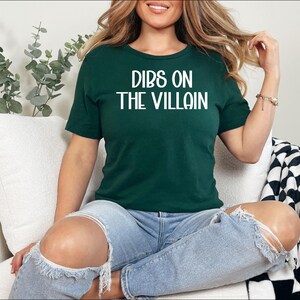 May include: A dark green t-shirt with the phrase "DIBS ON THE VILLAIN" in white block letters. The shirt is worn with blue jeans. The person is sitting on a white couch with a black and white checkered blanket.