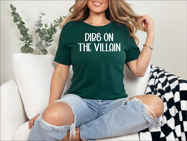 May include: A dark green t-shirt with the phrase "DIBS ON THE VILLAIN" in white block letters. The shirt is worn with blue jeans. The person is sitting on a white couch with a black and white checkered blanket.