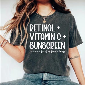 May include: Dark gray t-shirt with the text "RETINOL + VITAMIN C + SUNSCREEN" in white. Below the text is "these are a few of my favorite things". The shirt is worn by a person with long, wavy hair.