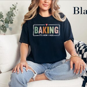 May include: A black short-sleeve t-shirt featuring a graphic design with the text BAKING in large, multi-colored block letters, a small red heart above, and the text IT'S HOW I HUG below, all enclosed within a thin white rectangular frame.