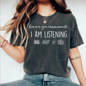 May include: Dark gray t-shirt with the text "however you communicate, I AM LISTENING" in white. The shirt features keyboard and sign language graphics. A casual, comfortable tee.