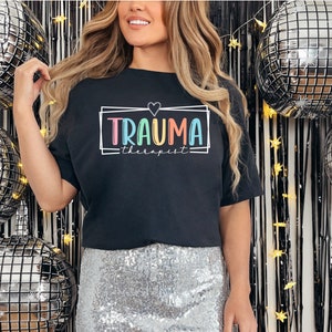 May include: Black t-shirt with a rainbow colored graphic that says "Trauma Therapist" in a box with a heart above the text.