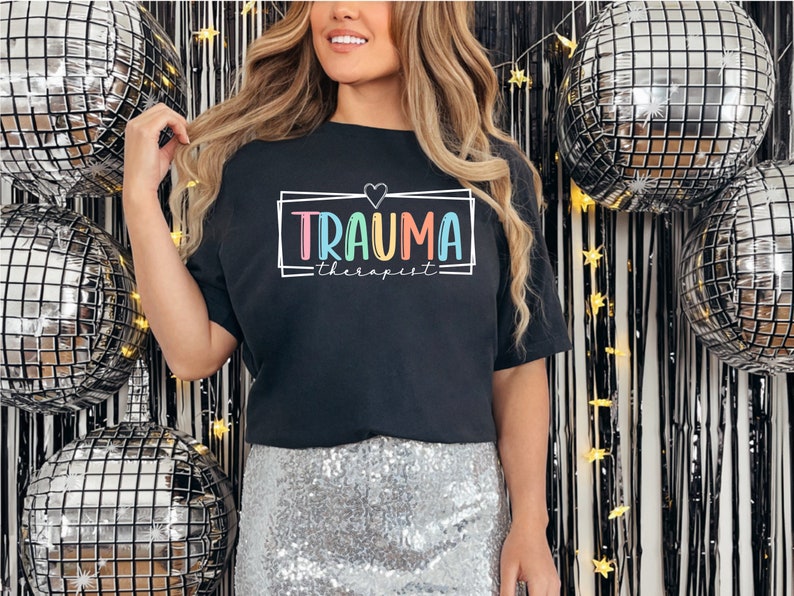 May include: Black t-shirt with a rainbow colored graphic that says "Trauma Therapist" in a box with a heart above the text.