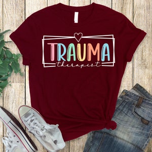 May include: A maroon t-shirt with a white graphic that says "Trauma Therapist" in colorful letters with a heart above the text.