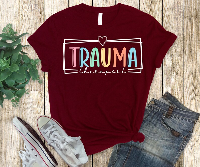 May include: A maroon t-shirt with a white graphic that says "Trauma Therapist" in colorful letters with a heart above the text.