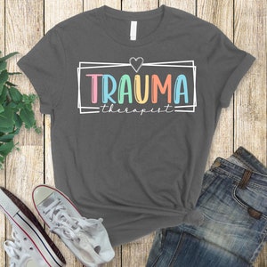 May include: A dark gray t-shirt with a white box around the text "TRAUMA therapist" in rainbow colors. A small white heart is above the text.