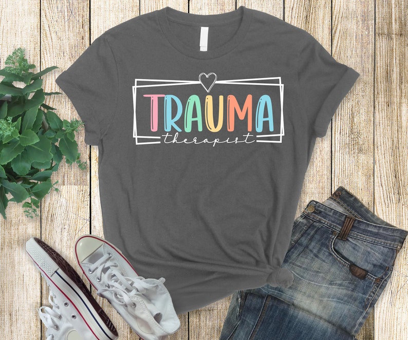 May include: A dark gray t-shirt with a white box around the text "TRAUMA therapist" in rainbow colors. A small white heart is above the text.