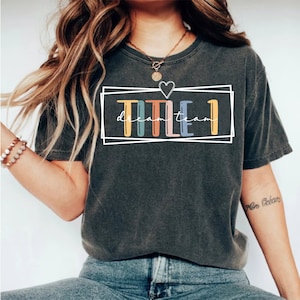 May include: A dark gray t-shirt with a white outline of a box around the text "TITLE I dream team". The text is in rainbow colors.