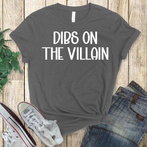 May include: A gray t-shirt with the text "DIBS ON THE VILLAIN" in white block letters. The shirt is paired with blue denim jeans and white sneakers with red accents. The items are arranged on a wooden surface.