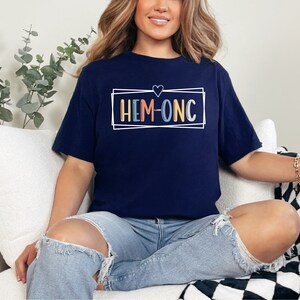 Puede incluir: Una camiseta azul marino con un cuadro blanco alrededor del texto "HEM-ONC" en letras de colores. Un peque&ntilde;o coraz&oacute;n est&aacute; por encima del texto.