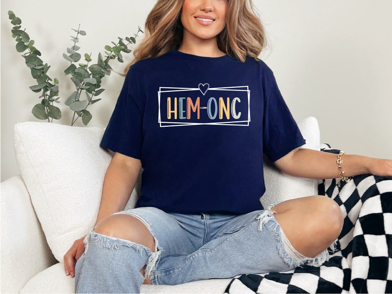 Puede incluir: Una camiseta azul marino con un cuadro blanco alrededor del texto "HEM-ONC" en letras de colores. Un peque&ntilde;o coraz&oacute;n est&aacute; por encima del texto.