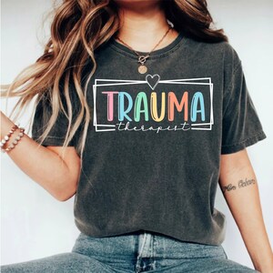 May include: A dark gray t-shirt with the text "TRAUMA therapist" in colorful letters inside a white box with a heart above the text.
