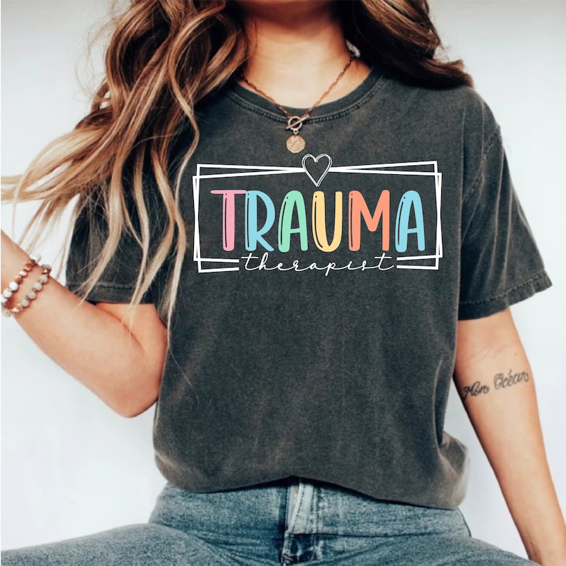 May include: A dark gray t-shirt with the text "TRAUMA therapist" in colorful letters inside a white box with a heart above the text.