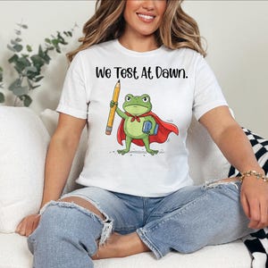 May include: White t-shirt with the text "We Test At Dawn." The shirt features a cartoon frog wearing a cape and holding a pencil and a book. The frog is green with a red cape.