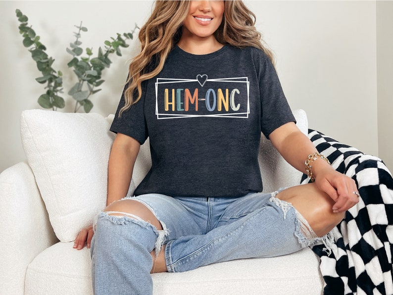 Puede incluir: Una camiseta gris oscuro con un contorno blanco de un coraz&oacute;n y un rect&aacute;ngulo alrededor del texto "HEM-ONC" en colores arco&iacute;ris.
