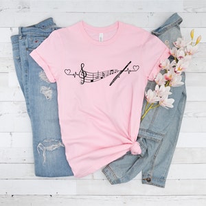 Może przedstawiać: R&oacute;żowy T-shirt z czarnym nadrukiem przedstawiającym nutę muzyczną i flet z linią bicia serca i sercami po obu stronach. Koszulka leży na białym drewnianym tle z niebieskimi dżinsami i dżinsową kurtką z gałązką białych kwiat&oacute;w.
