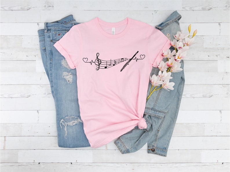 Może przedstawiać: R&oacute;żowy T-shirt z czarnym nadrukiem przedstawiającym nutę muzyczną i flet z linią bicia serca i sercami po obu stronach. Koszulka leży na białym drewnianym tle z niebieskimi dżinsami i dżinsową kurtką z gałązką białych kwiat&oacute;w.